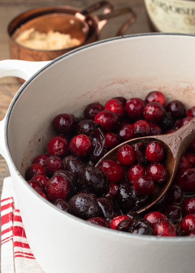 cranberries and frozen dark cherries starting to cook down in a white pot with a wooden spoon