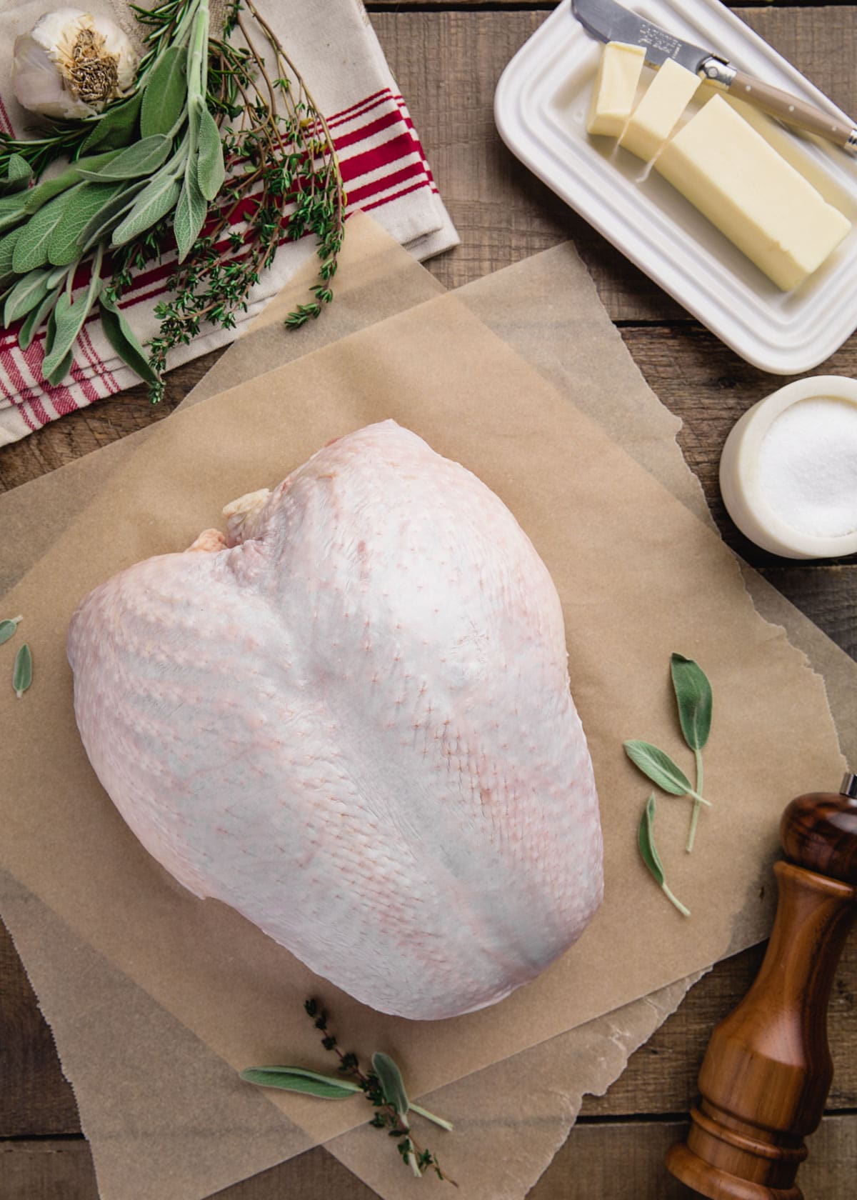 raw whole turkey breast on parchment paper next to a stick of butter, herbs, and seasonings