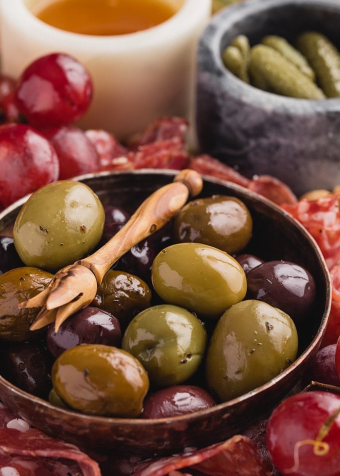 bowl of olives, honey, and cornichons with charcuterie and fruit on a cheese board