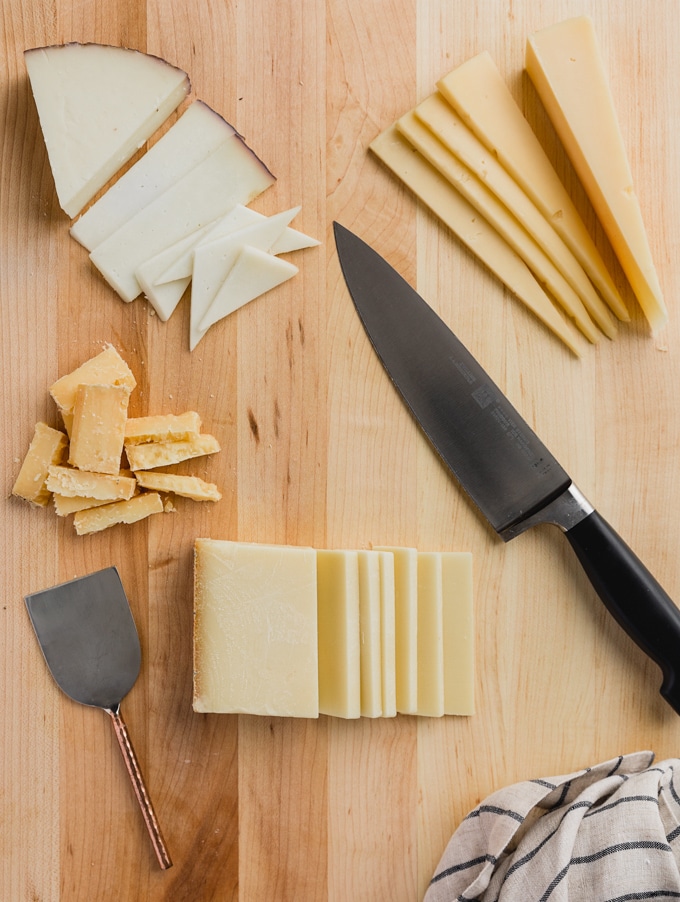 sliced blocks of cheese on a wood cutting board with a chef's knife and a cheese knife