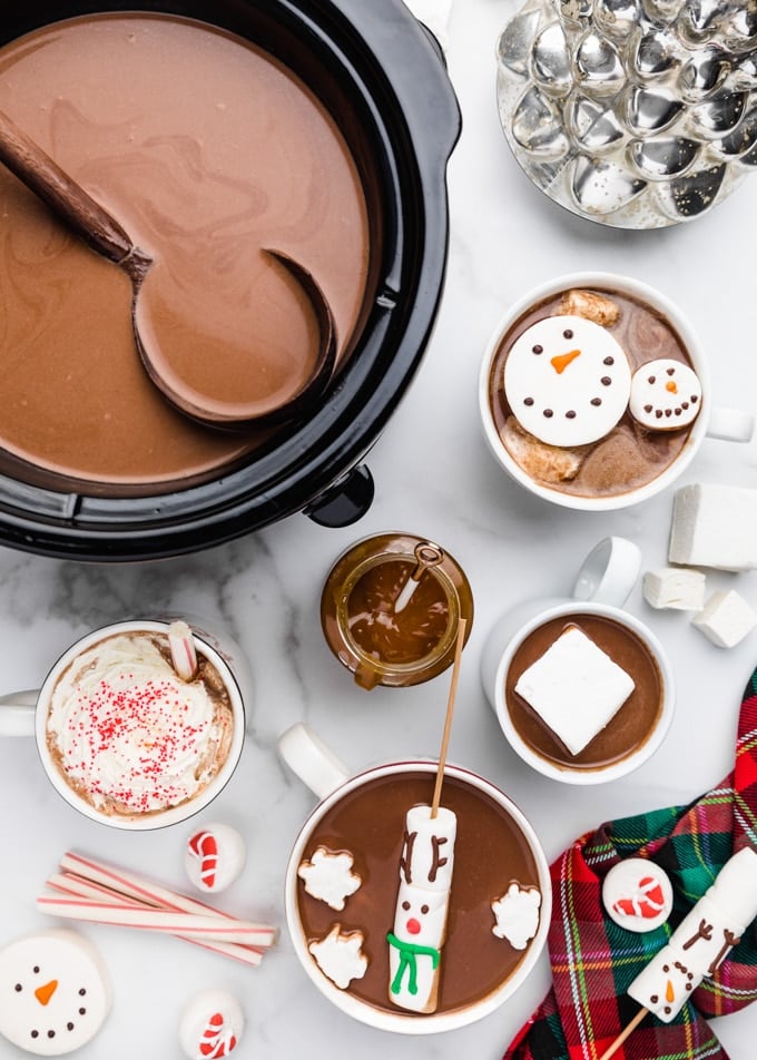 hot chocolate in a crockpot with a ladle, surrounded buy mugs of hot chocolate with snowman and reindeer marshmallows