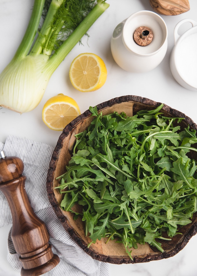 overhead shot of a whole bulb of fennel, lemon halves, a bowl of baby arugula, olive oil, salt, and pepper on white marble