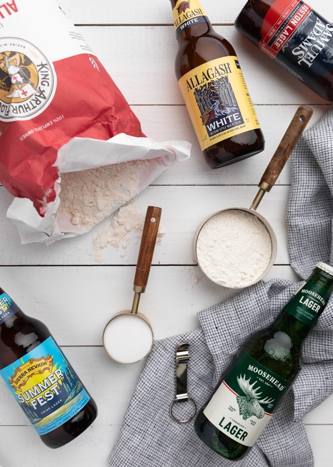 flatlay showing an open bag of all purpose flour, measuring cups filled with flour and sugar, and 5 different bottles of beer