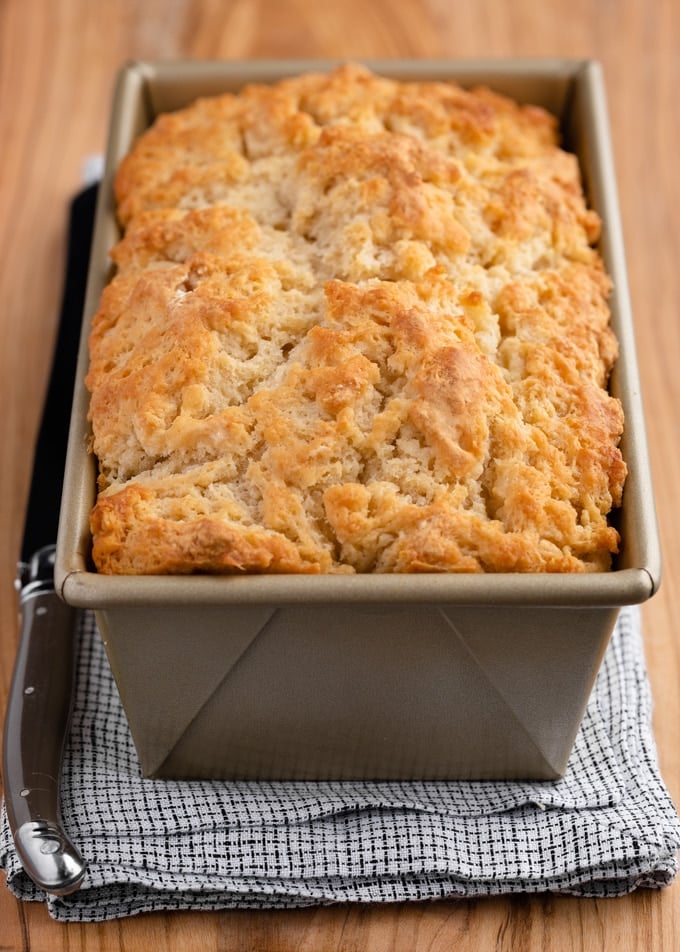 baked loaf of beer bread in a gold loaf pan on a wooden board with a white and blue checked napkin