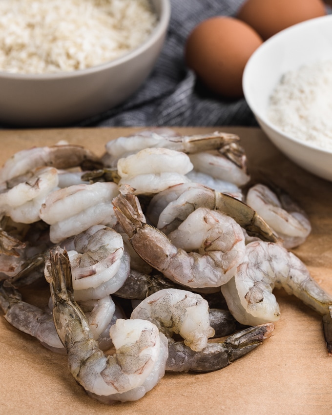 raw extra jumbo shrimp on parchment paper alongside bowls of breading ingredients