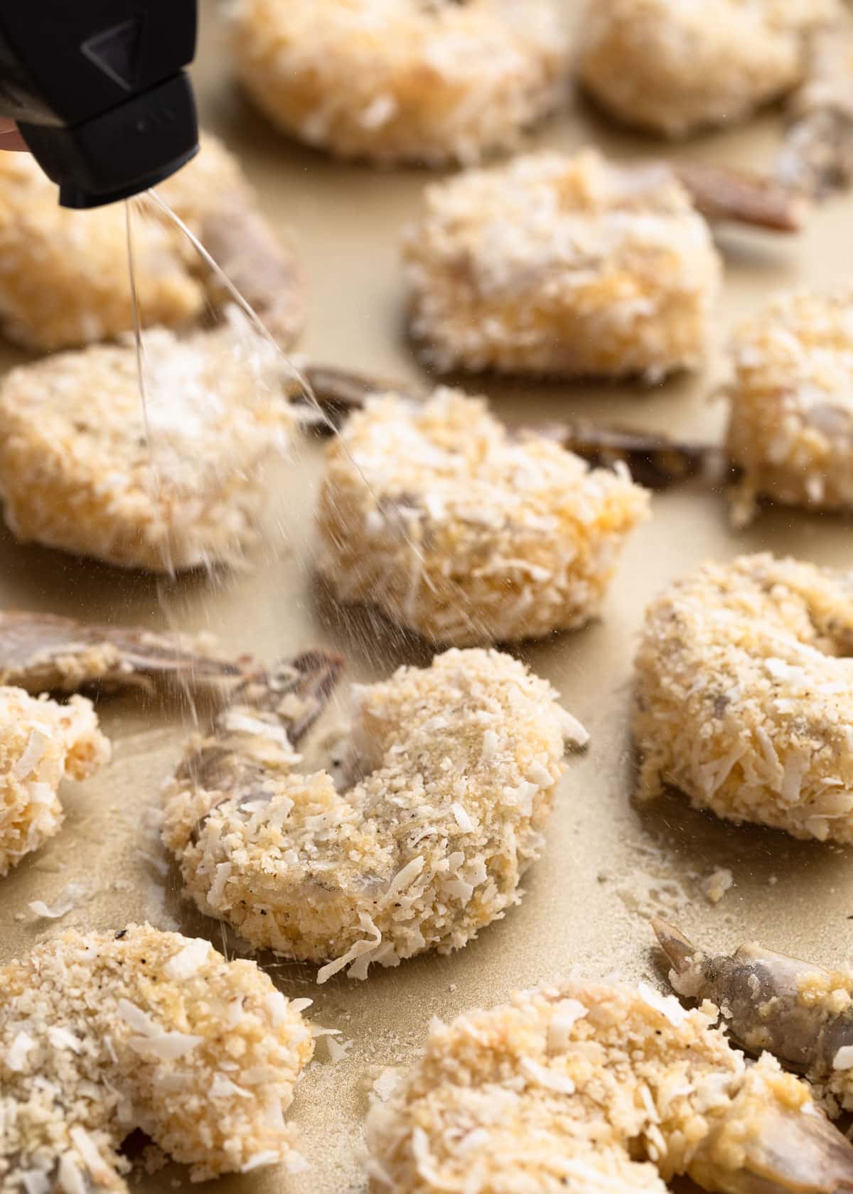 raw coconut shrimp on a baking sheet being sprayed with oil from a mister bottle