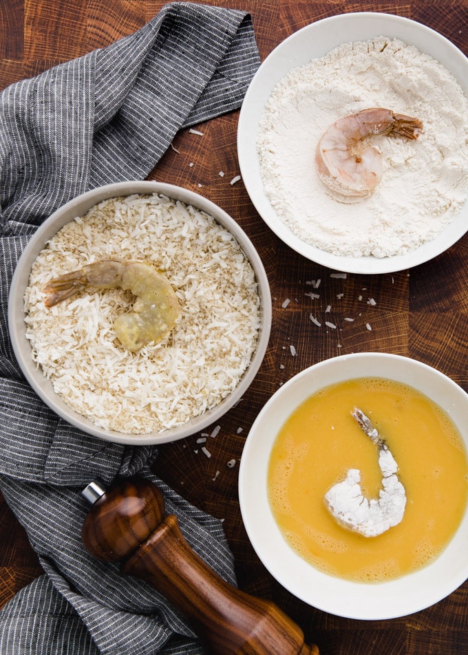 three bowls showing the process of breading coconut shrimp: eggs wash, flour, and a coconut-panko mixture