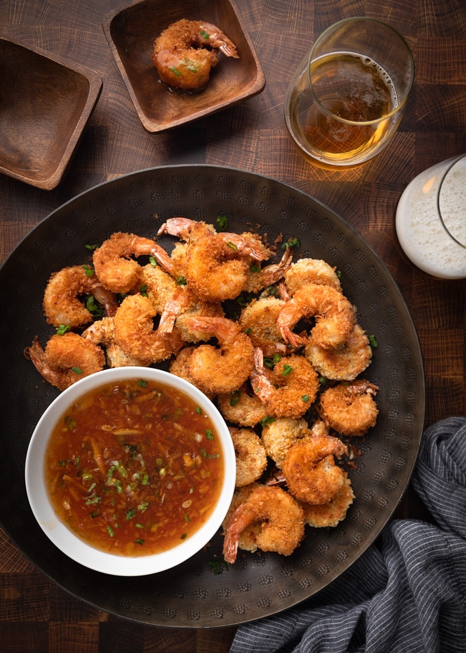 coconut shrimp on a dark bronze serving platter with orange dipping sauce, next to two glasses of beer