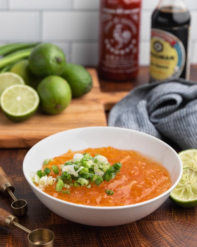 orange marmalade in a white bowl with sliced scallions against a background of whole and cut limes, and bottles of soy sauce and sriracha