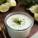 cilantro lime sauce in a glass jar on a wood board, with bunches of fresh cilantro and fresh limes