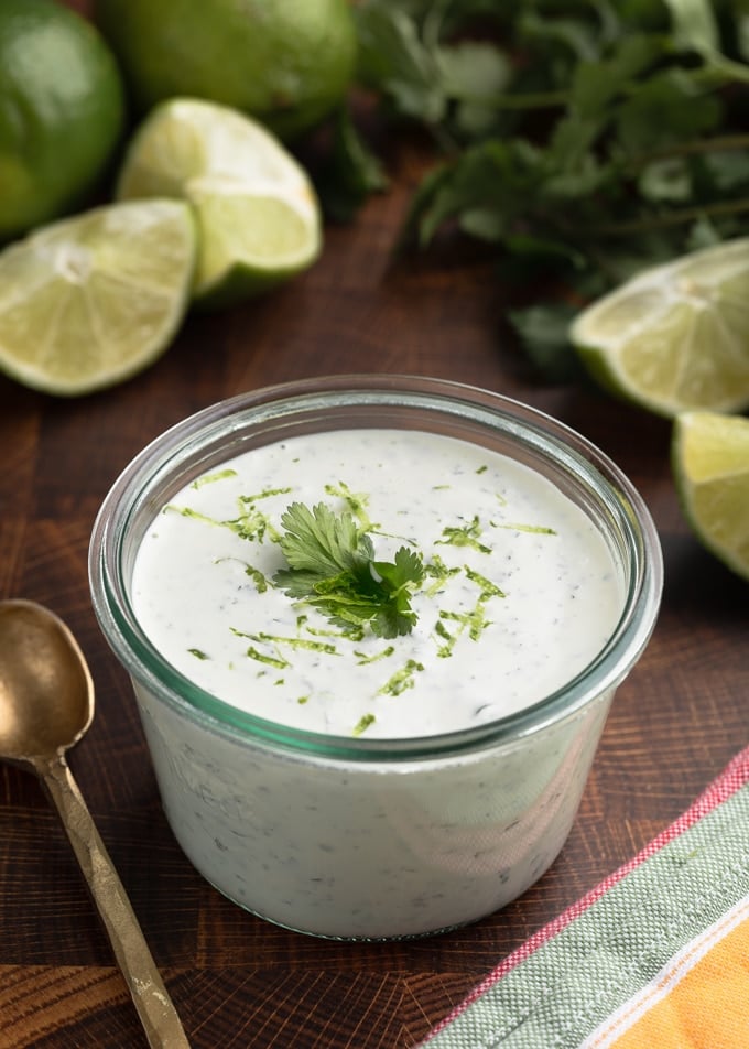 cilantro lime sauce in a glass jar on a wood board, with bunches of fresh cilantro and fresh limes