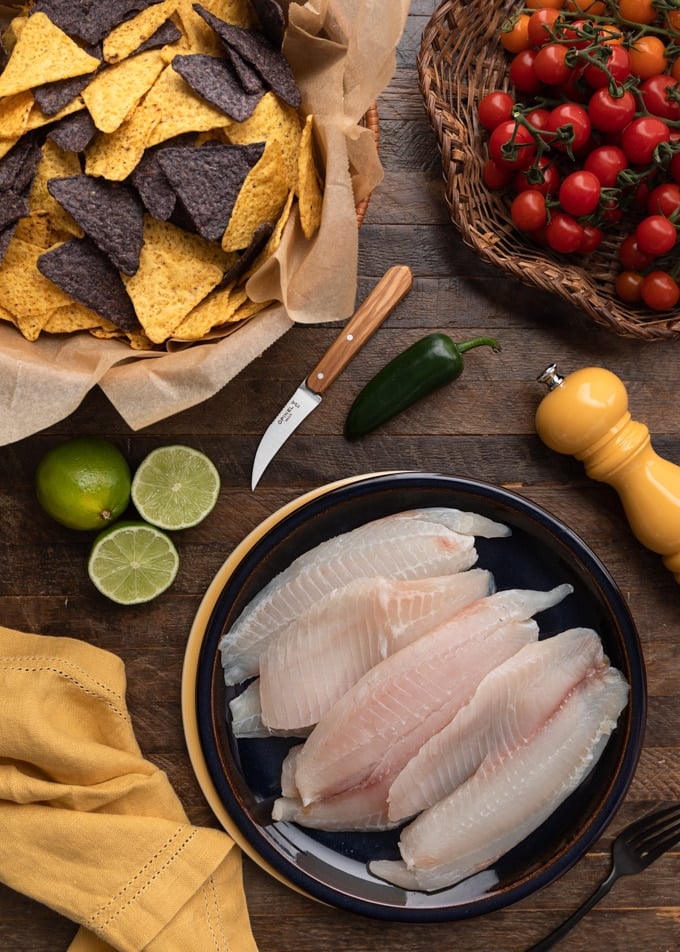 raw tilapia fillets on a plate next to a baskets of tortilla chips and cherry tomatoes on the vine