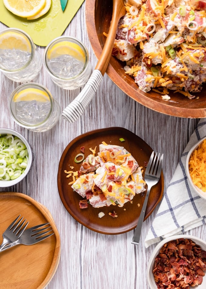 overhead photo of bacon cheddar potato salad in a wooden serving bowl, with a portion on a wooden plate and glasses of cold summer beverages