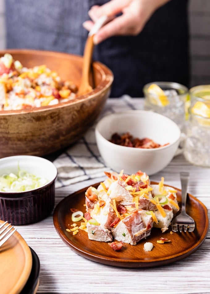 loaded potato salad with bacon and cheddar on a wooden plate, surrounded by bowls of scallions, bacon, and a large serving bowl