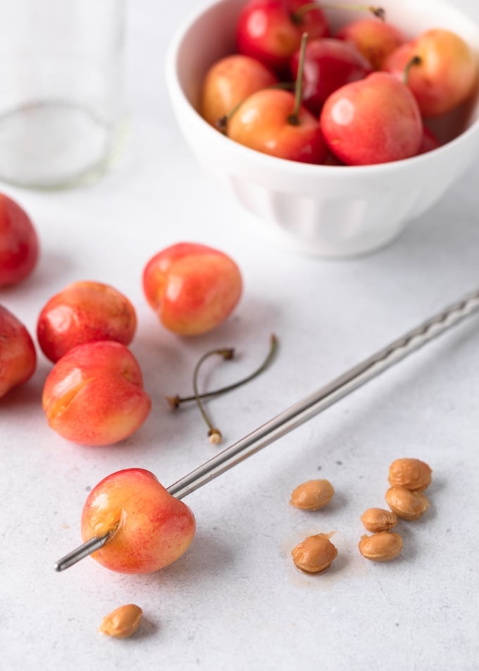 A Rainier cherry skewered with a metal chopstick next to a pile of cherry pits, pitted cherries, and a bowl of whole cherries