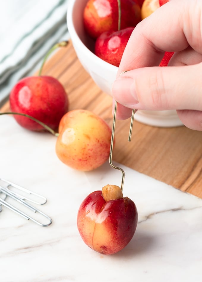 Pit being removed from a Rainier cherry with a gold-tone paper clip, on a marble cutting board
