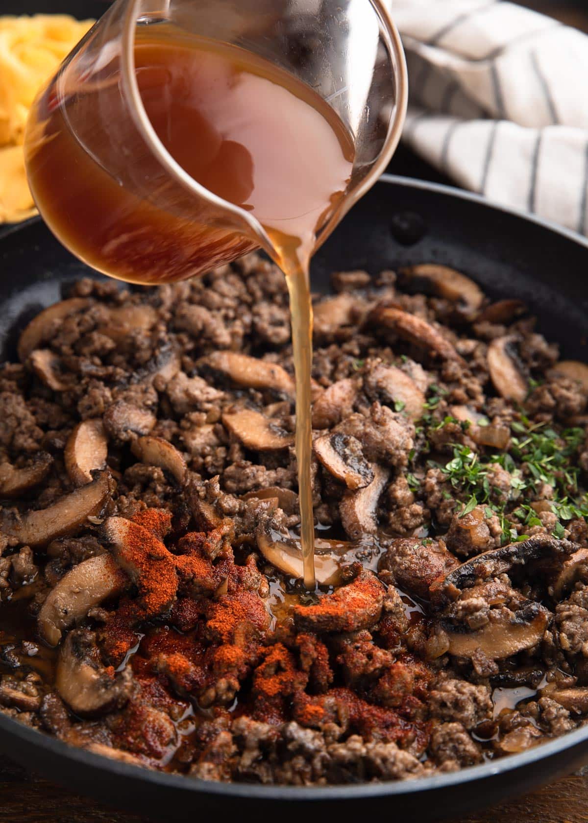 pouring beef broth into a saute pan with ground beef, mushrooms, paprika, and herbs