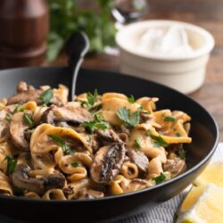 ground beef stroganoff in a black ceramic bowl with a black fork, in front of a bowl of sour cream, a glass of red wine, and a pepper mill