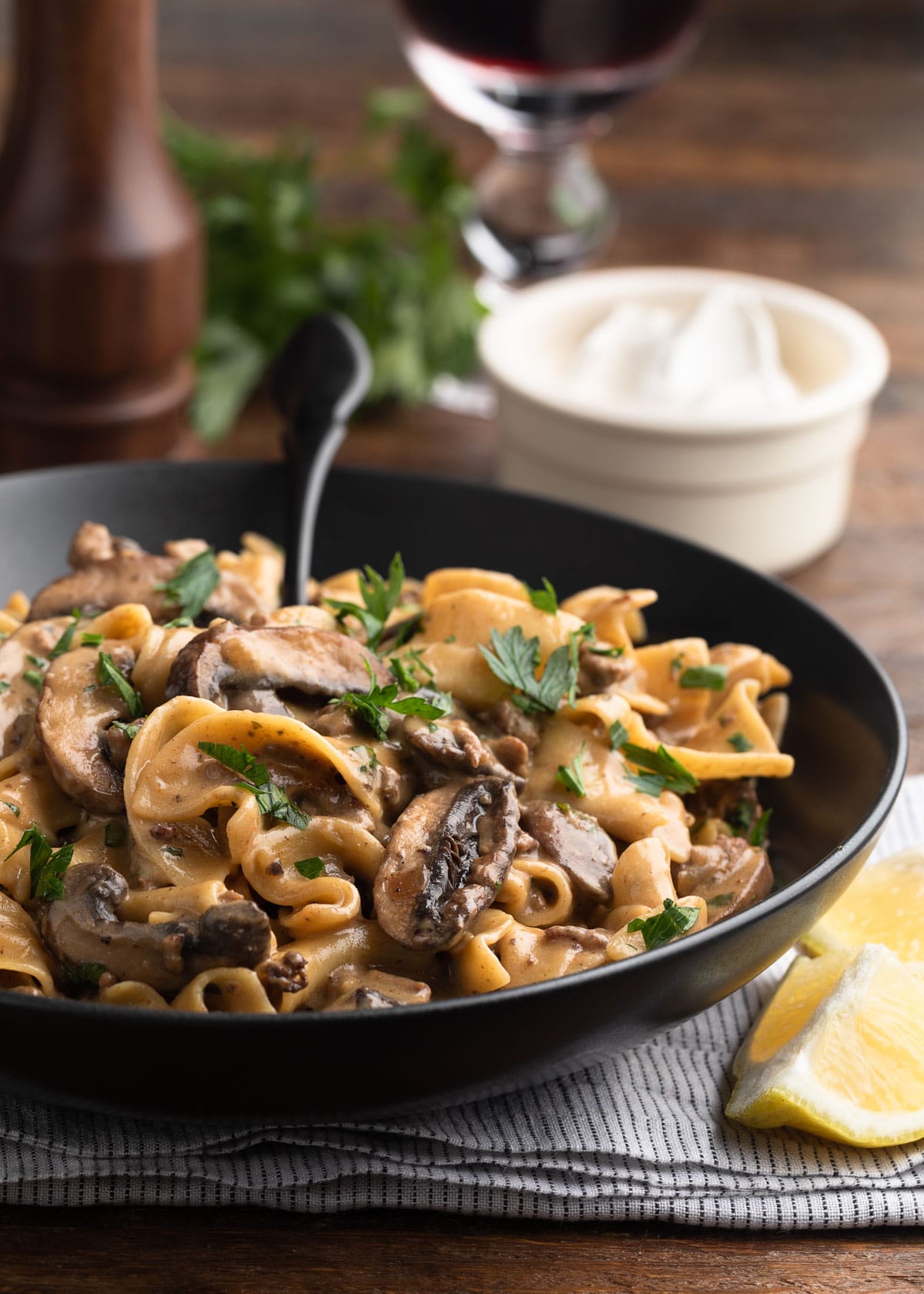 ground beef stroganoff in a black ceramic bowl with a black fork, in front of a bowl of sour cream, a glass of red wine, and a pepper mill