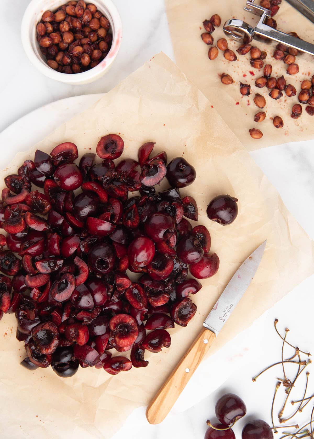 chopped cherries on a parchment-lined marble board, with a paring knife, cherry pitting tool, and bowl of cherry pits