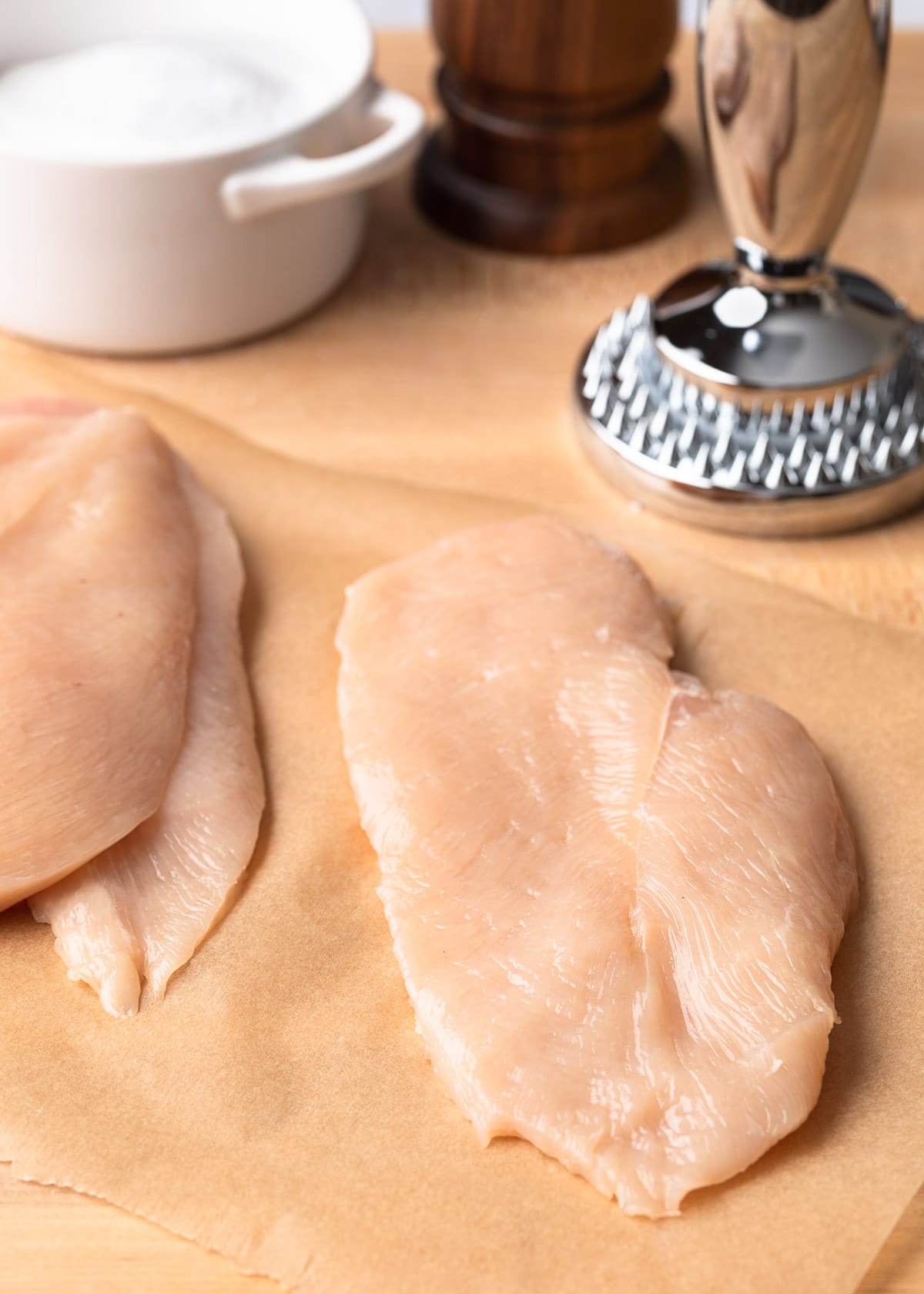 thin sliced chicken cutlets on parchment paper next to a stainless steel meat pounder, salt crock, and pepper grinder.