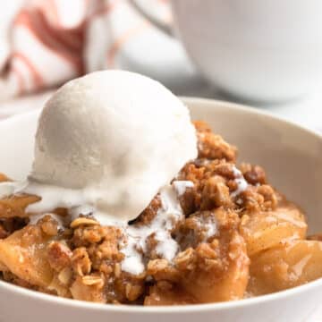 apple crisp in a white bowl with a scoop of vanilla ice cream and a cup of tea in the background