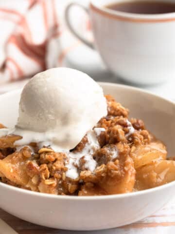 apple crisp in a white bowl with a scoop of vanilla ice cream and a cup of tea in the background