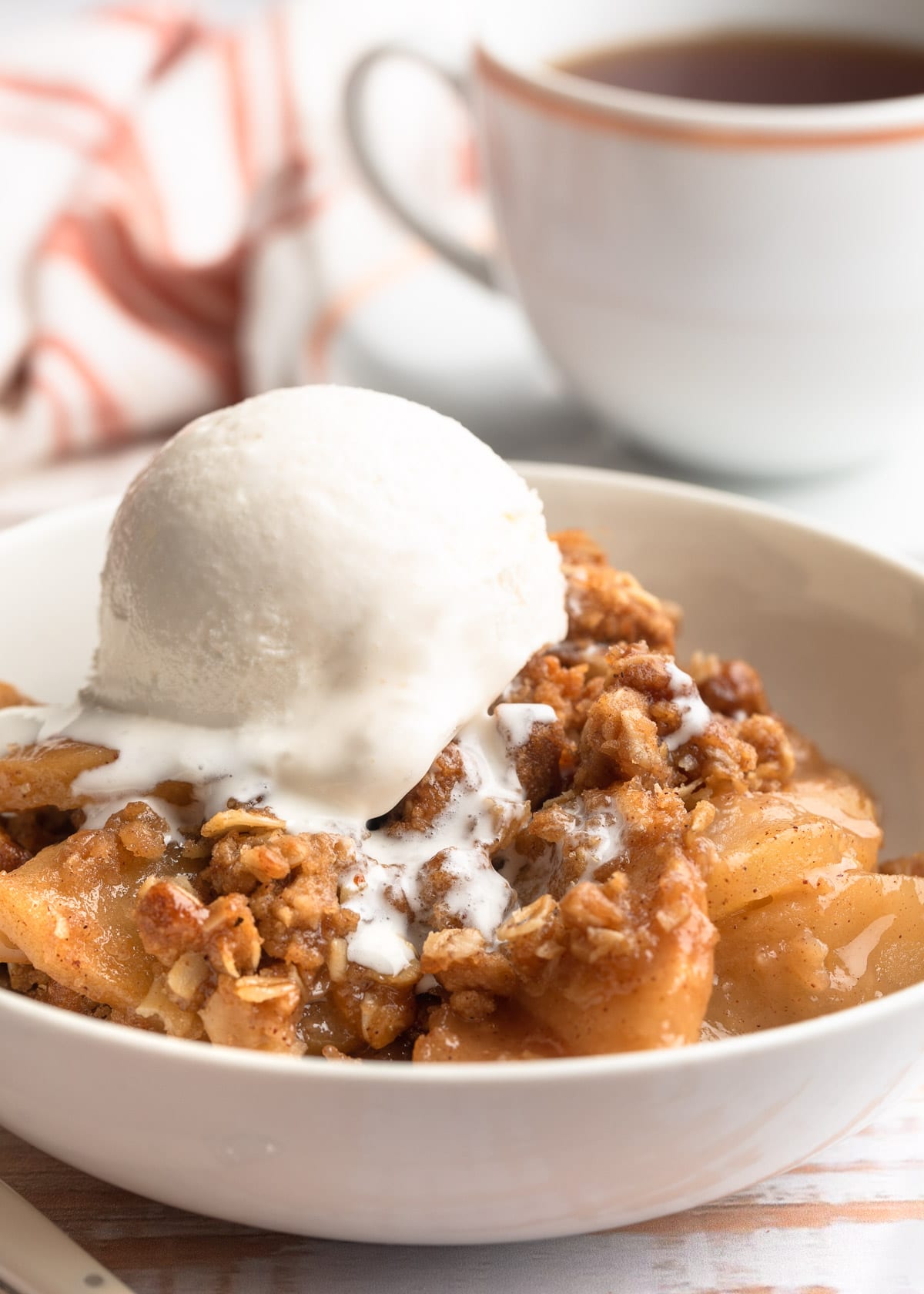 apple crisp in a white bowl with a scoop of vanilla ice cream and a cup of tea in the background