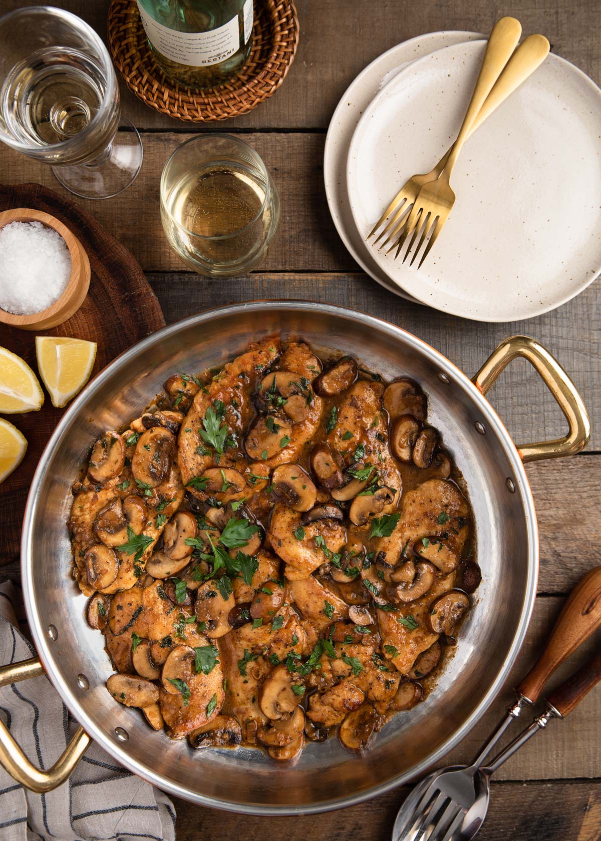 overhead of chicken marsala in a copper pan on a wood board with serving plates and glasses of wine