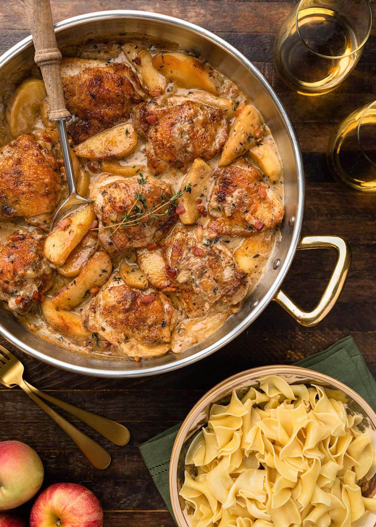 chicken thighs and apples in a copper frying pan with hard cider sauce next to a bowl of egg noodles