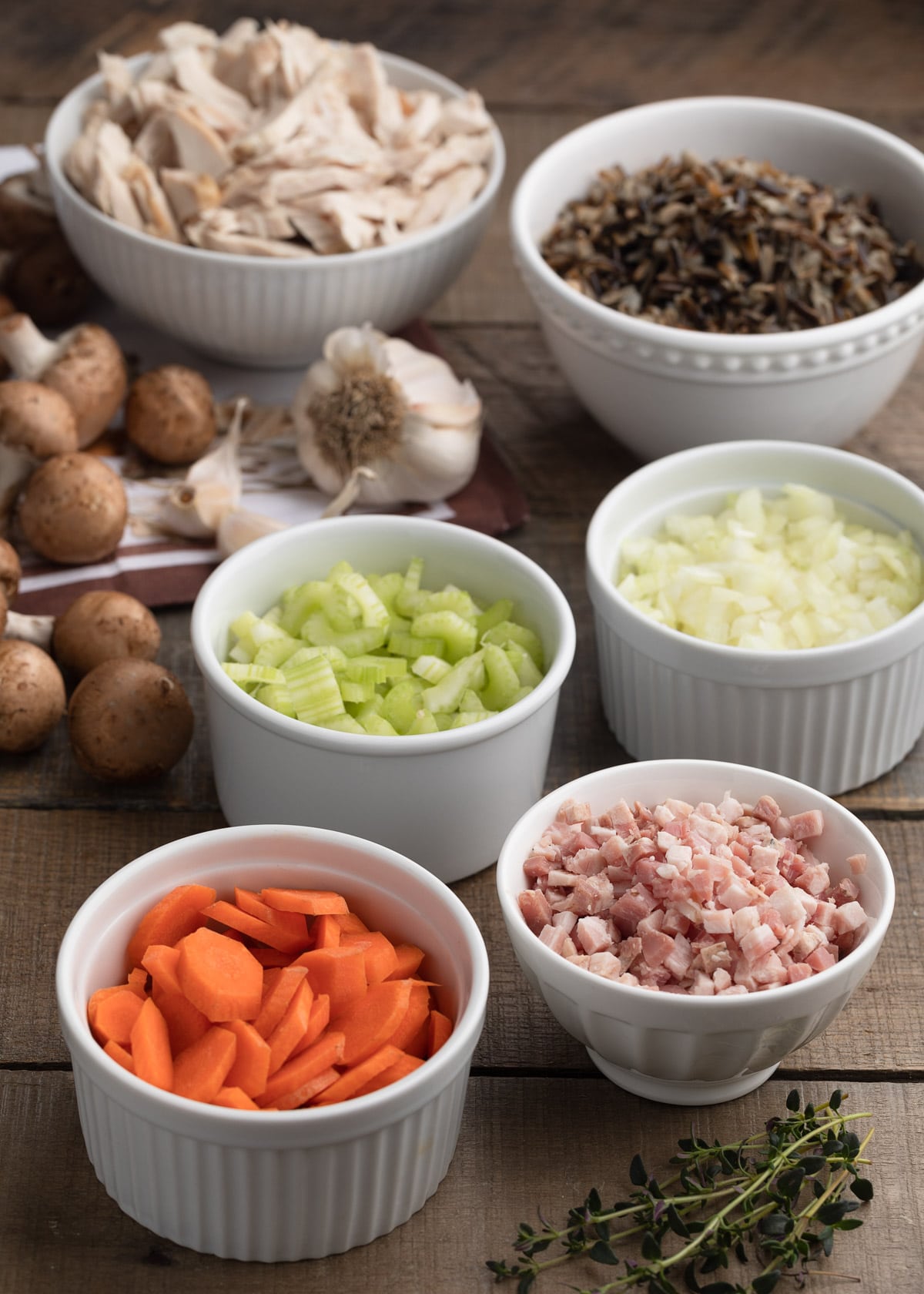 bowls of sliced carrots, pancetta, celery, chopped onions, cooked wild rice, and shredded turkey breast on a wood board, with mushrooms, herbs, and garlic cloves