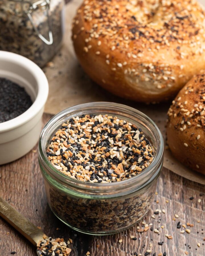 everything bagel seasoning in a glass jar, surrounded by everything bagels on a wood board