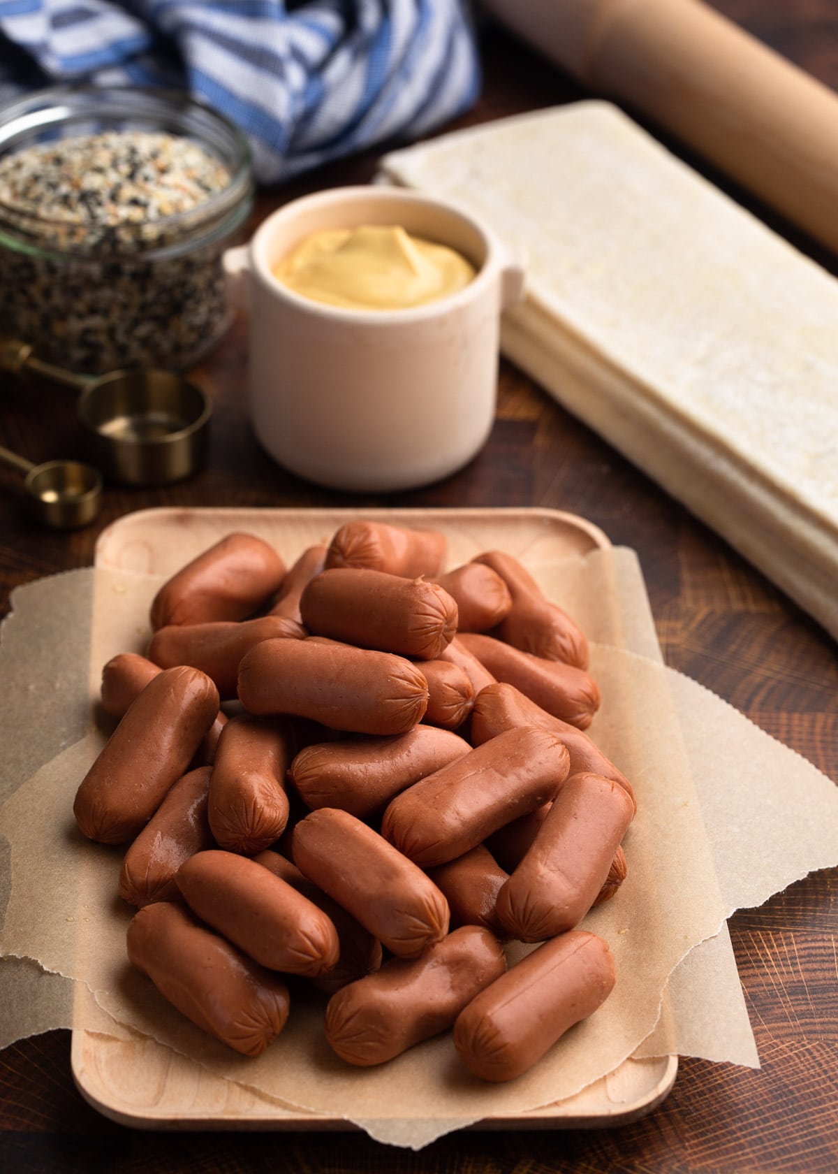 beef cocktail franks on a wooden board, surrounded by sheets of puff pastry, a crock of dijon, and a jar of seasoning