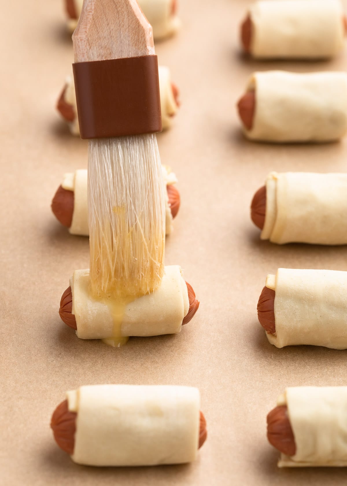 brushing uncooked pigs in a blanket with egg wash on a baking sheet