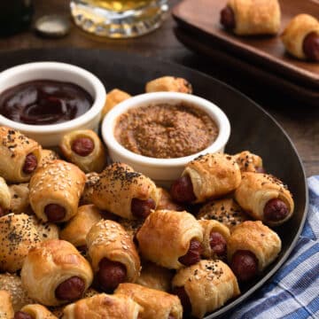 puff pastry pigs in a blanket on a bronze serving platter with bowls of grainy mustard and bbq sauce