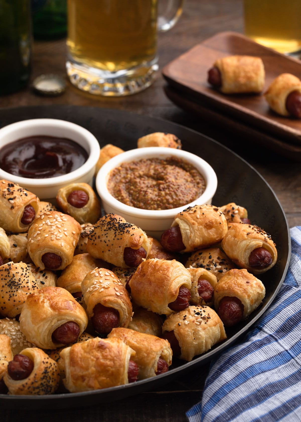 puff pastry pigs in a blanket on a bronze serving platter with bowls of grainy mustard and bbq sauce