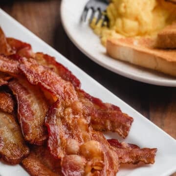  bacon cuit au four sur un plateau blanc devant une assiette de petit-déjeuner composée d'œufs brouillés et de pain grillé