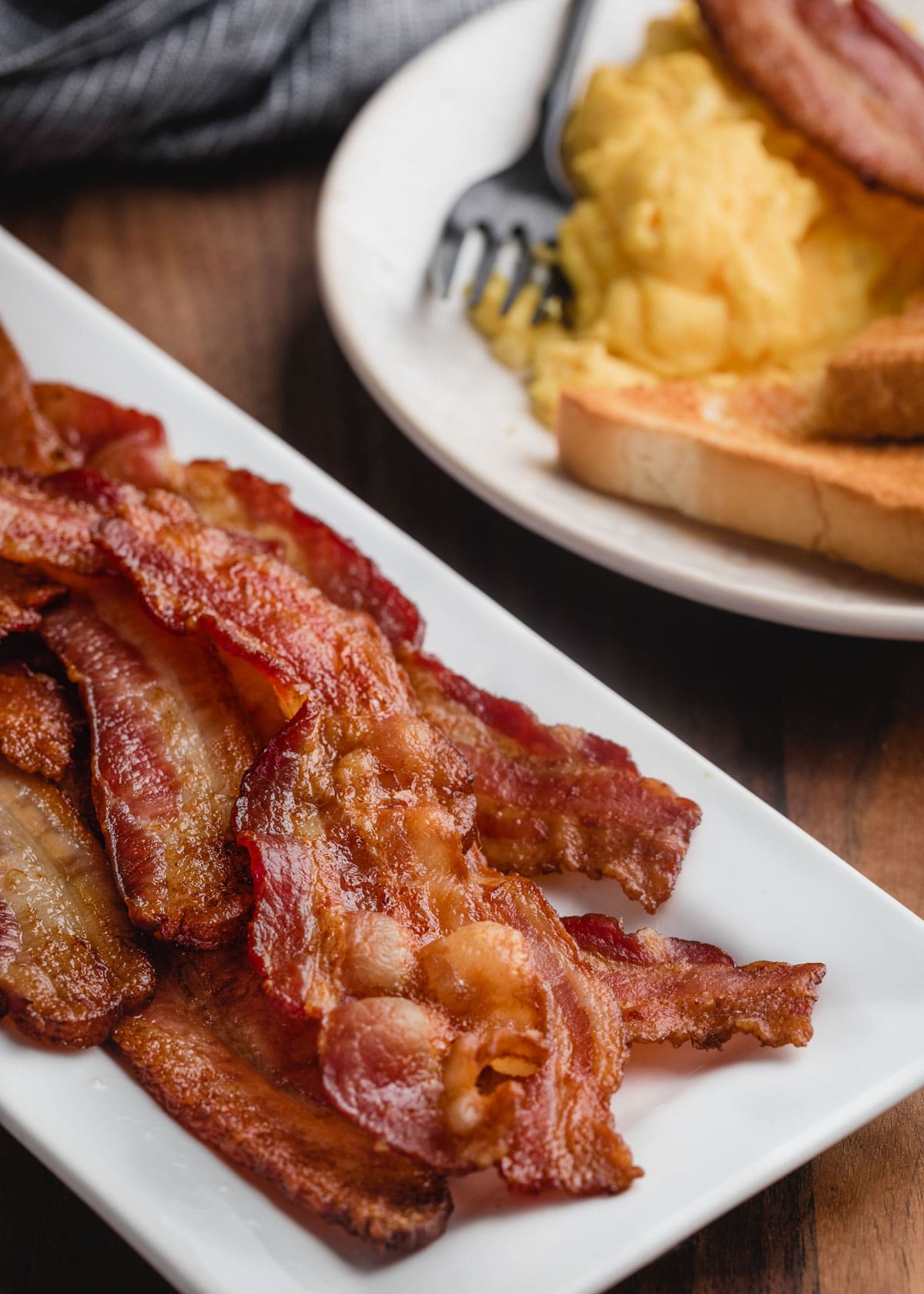 baked bacon on a white platter in front of a breakfast plate of scrambled eggs and toast