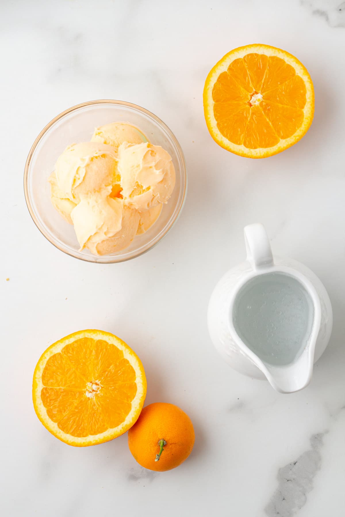 overhead photo of a bowl of orange sherbet, orange halves, and a pitcher of sparkling water
