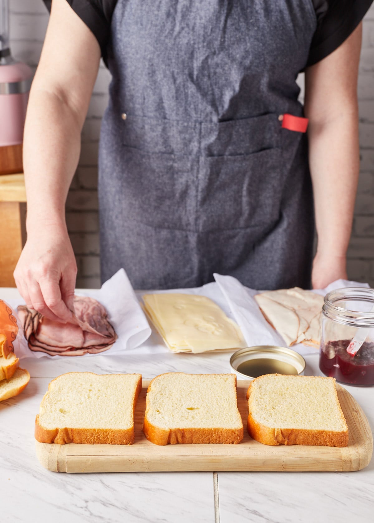 three slices of bread on a wood cutting board, in front of piles of deli meat and cheese, and a woman in an apron in the background picking up a slice of ham