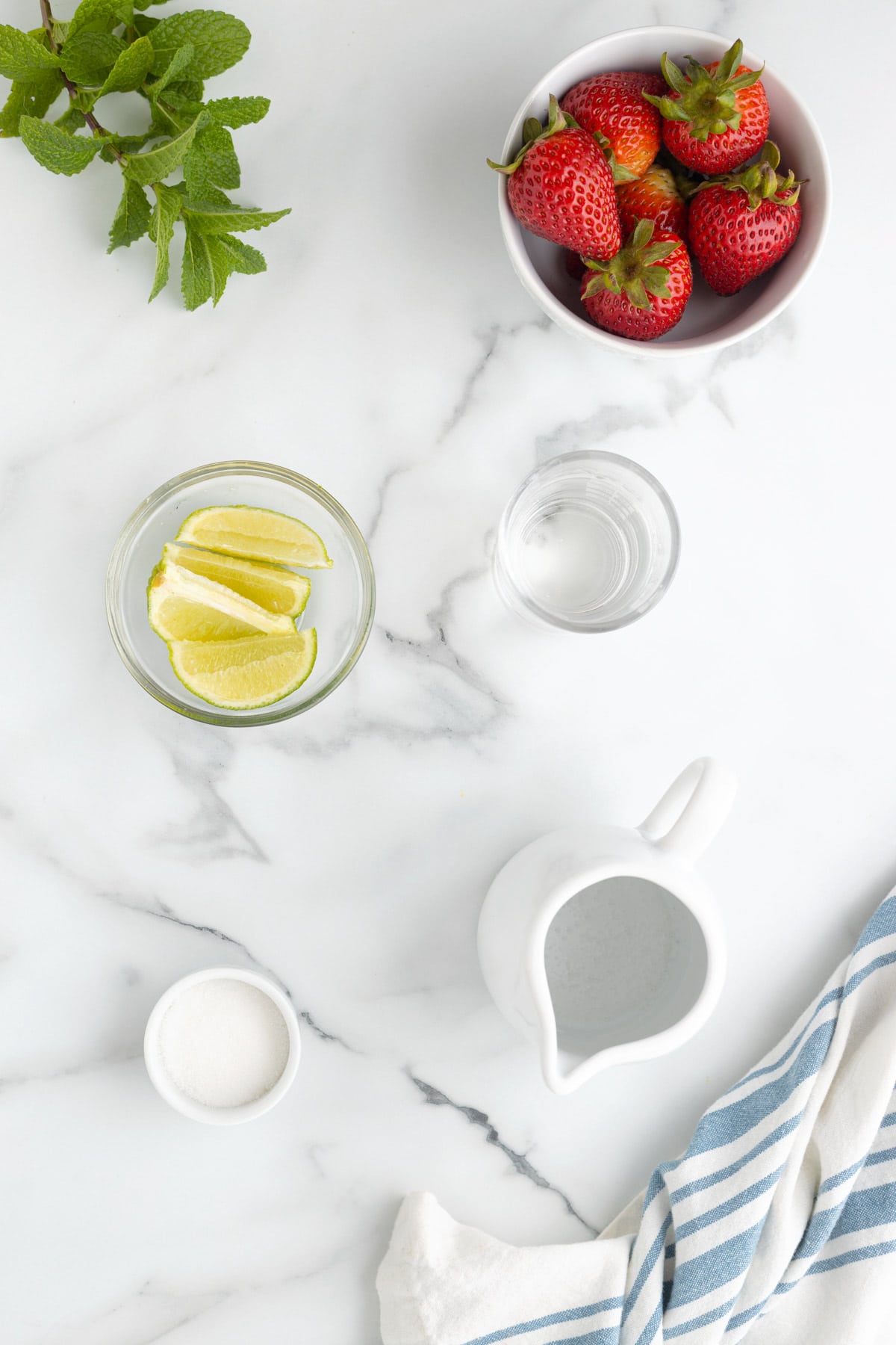 overhead of a bowl of strawberries, a bowl of lime wedges, sugar, rum, a sprig of mint, and sparkling water on a white marble background