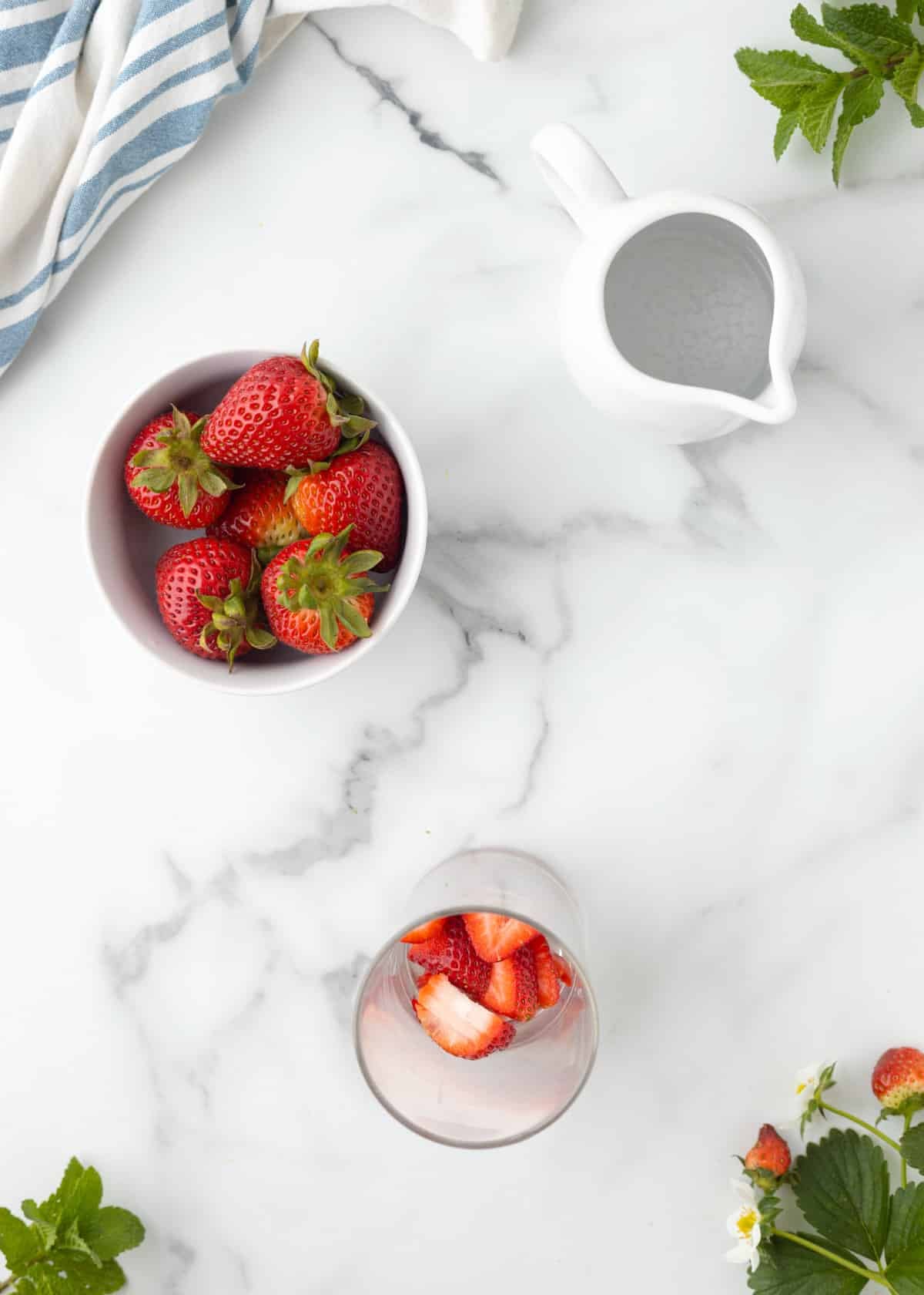 Overhead view of fresh strawberries in a bowl with sliced strawberries in a glass, alongside mint leaves and a small pitcher on a marble surface.