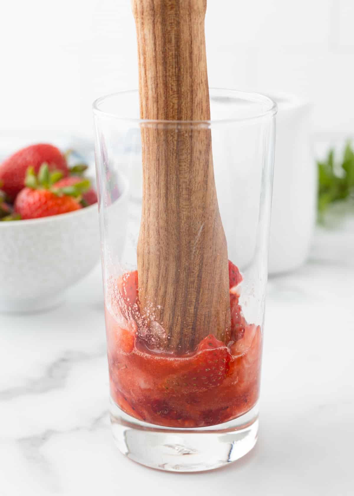 Muddled strawberries being crushed with a wooden muddler in a tall glass, releasing juices for a strawberry mojito.