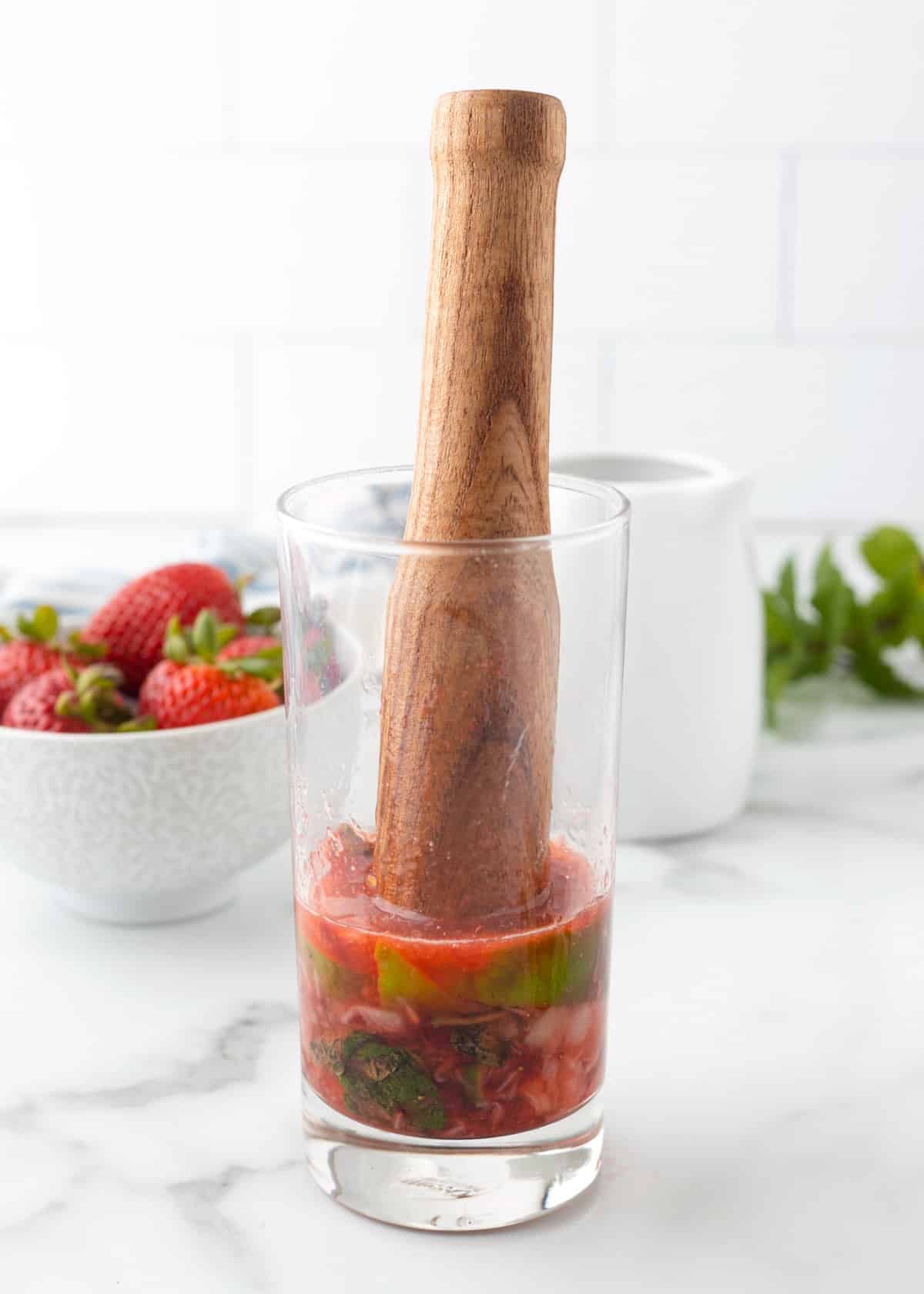 Muddled strawberries, lime, and mint being pressed with a wooden muddler in a tall glass, forming the base of a strawberry mojito.