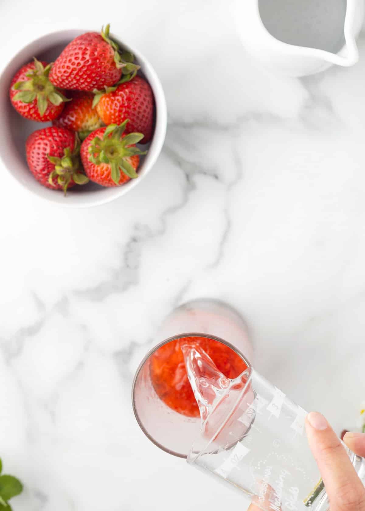 White rum being poured from a measuring glass into a tall glass of muddled strawberries, building the base for a strawberry mojito.