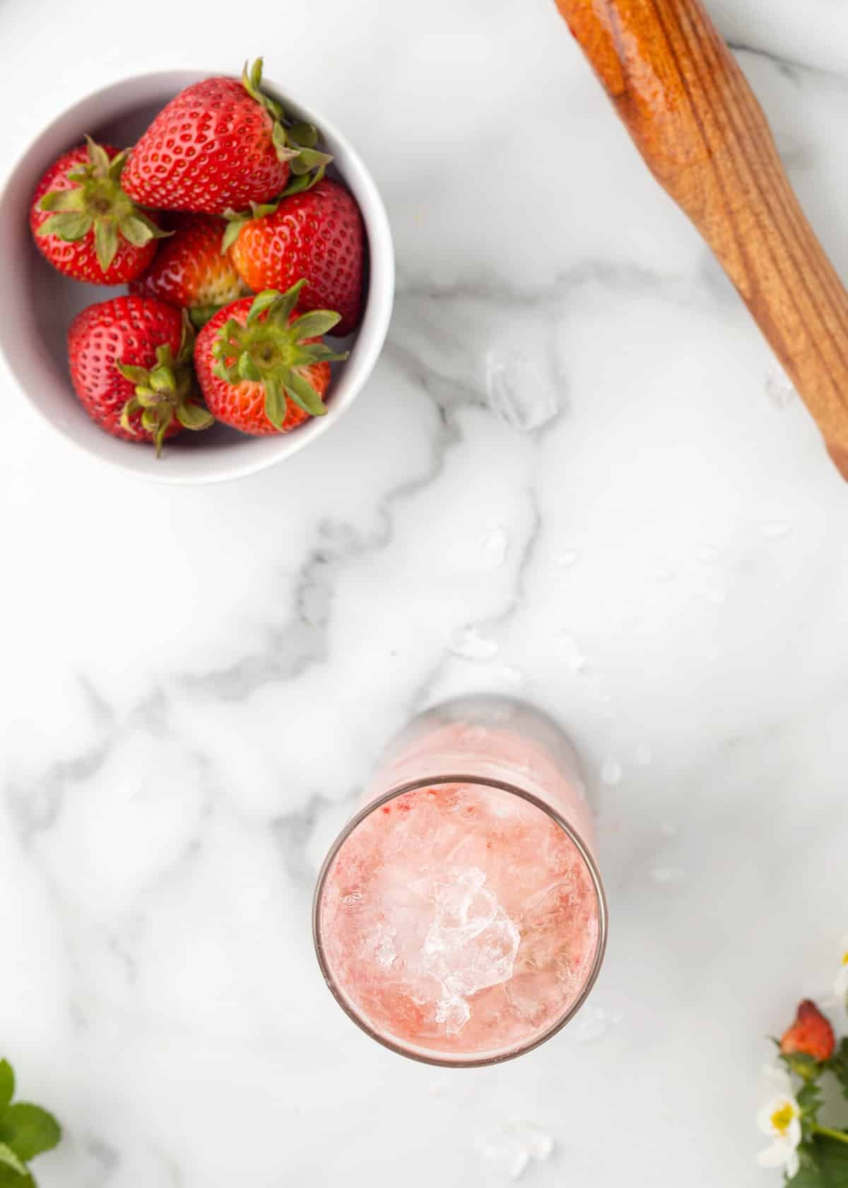 Top-down view of a strawberry mojito in progress with ice in the glass, a bowl of fresh strawberries nearby, and a wooden muddler on the marble surface.
