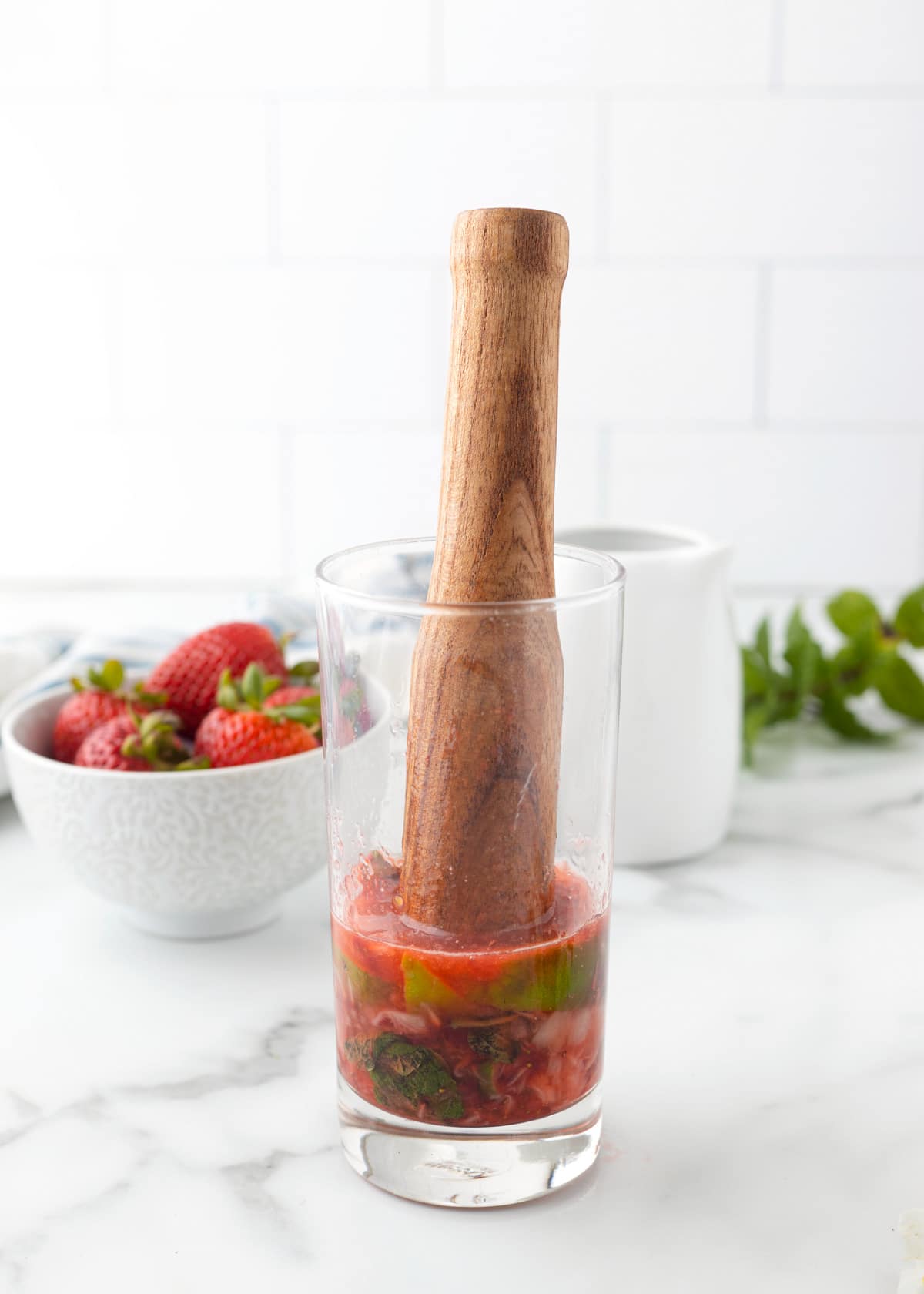 a wooden muddler in a highball glass with strawberries, lime wedges, and mint