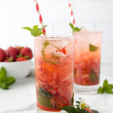 two strawberry mojitos in highball glasses with red and white paper straws and strawberry and herb garnishes