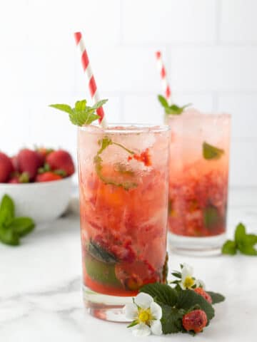two strawberry mojitos in highball glasses with red and white paper straws and strawberry and herb garnishes