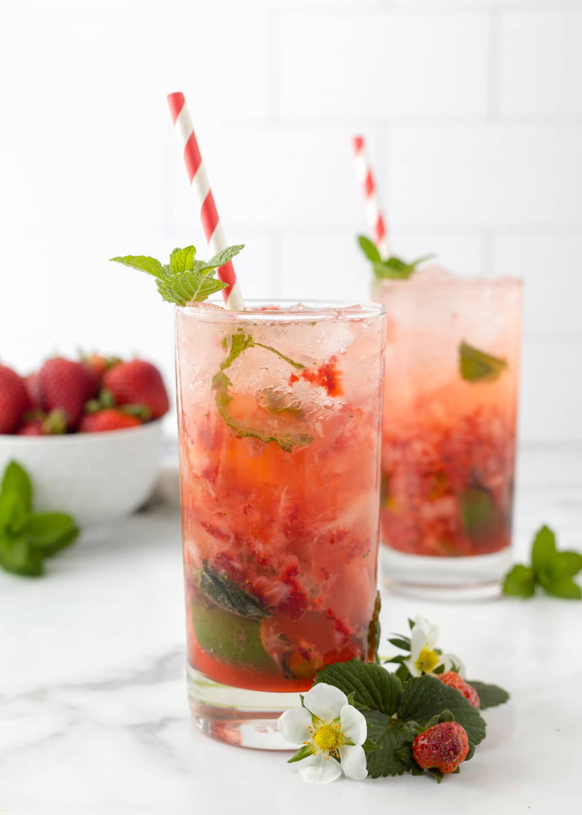 two strawberry mojitos in highball glasses with red and white paper straws and strawberry and herb garnishes